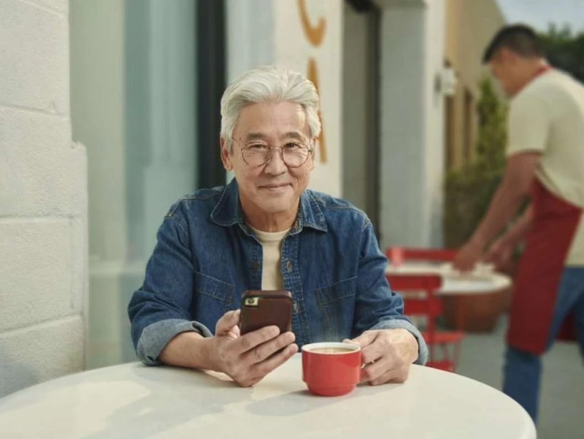 Elderly Asian man smiles while holding his smartphone and sits at a cafe table with a red mug and a waiter in the background.